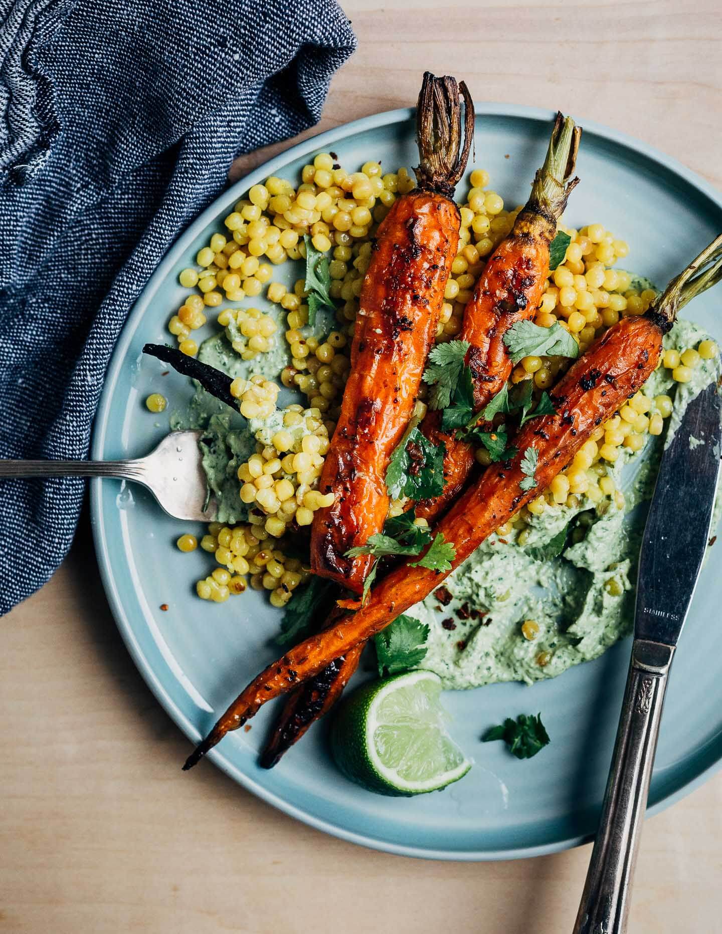 A close-up view of a plate with roasted carrots, green tahini, and couscous. A fork is prepped with a bite of food and there’s a knife alongside. 