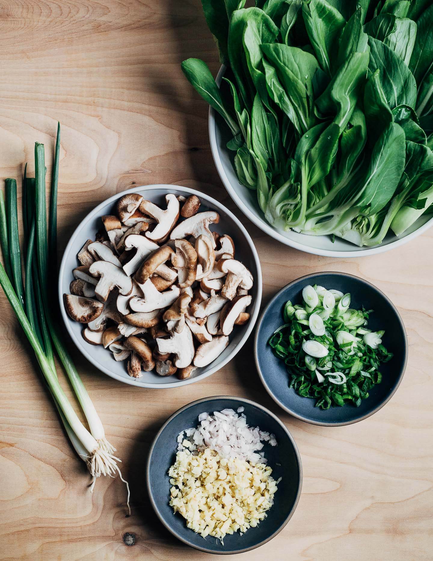 Bowls of bok choy, sliced mushrooms, ginger and garlic, and green onions on a tabletop. 