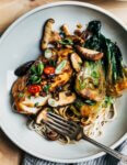 A plate with stir fried vegetables, noodles, and glazed cod. A fork is flaking of a bite of cod.