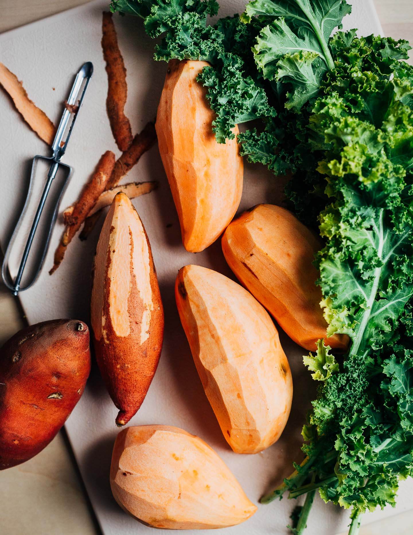 A cutting board with peeled sweet potatoes and leaves of kale. 