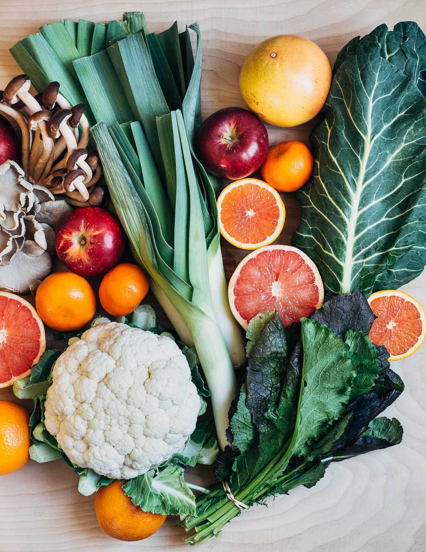 An array of produce on a tabletop. Includes citrus, cauliflower, greens, leeks, and apples. 