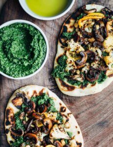 Two flatbread pizzas on a cutting board with a small bowl of pesto.