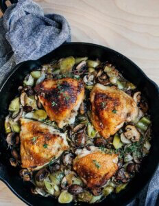 A cast iron skillet with chicken thighs, mushrooms, and leeks.