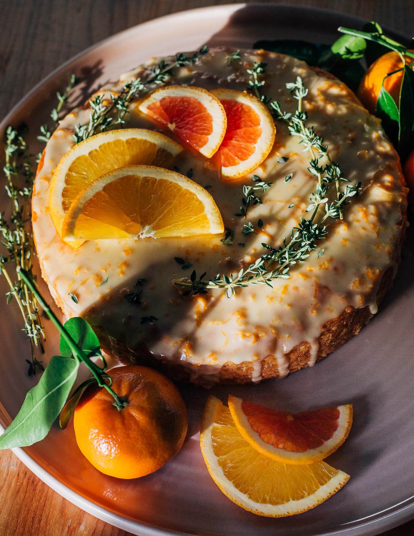 A platter with a glazed cake. It’s garnished with thyme sprigs, sliced citrus, and leafy whole citrus. Cake is pictured in raking sunlight.