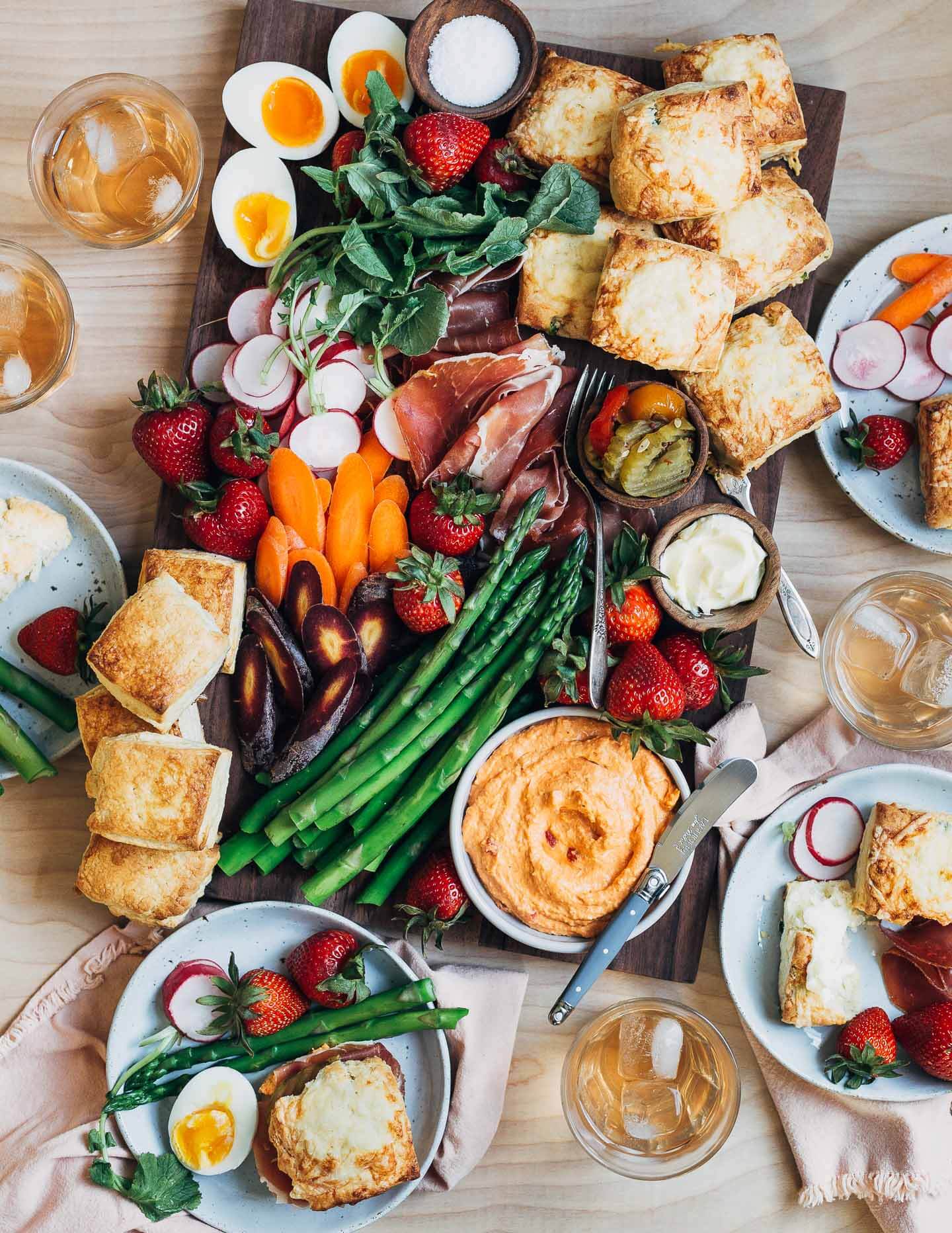 A crowded tabletop with a biscuit board, drinks, and little plates with food. 