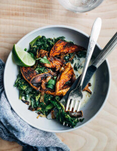 A bowl of roasted sweet potatoes with braised greens and a lime wedge.