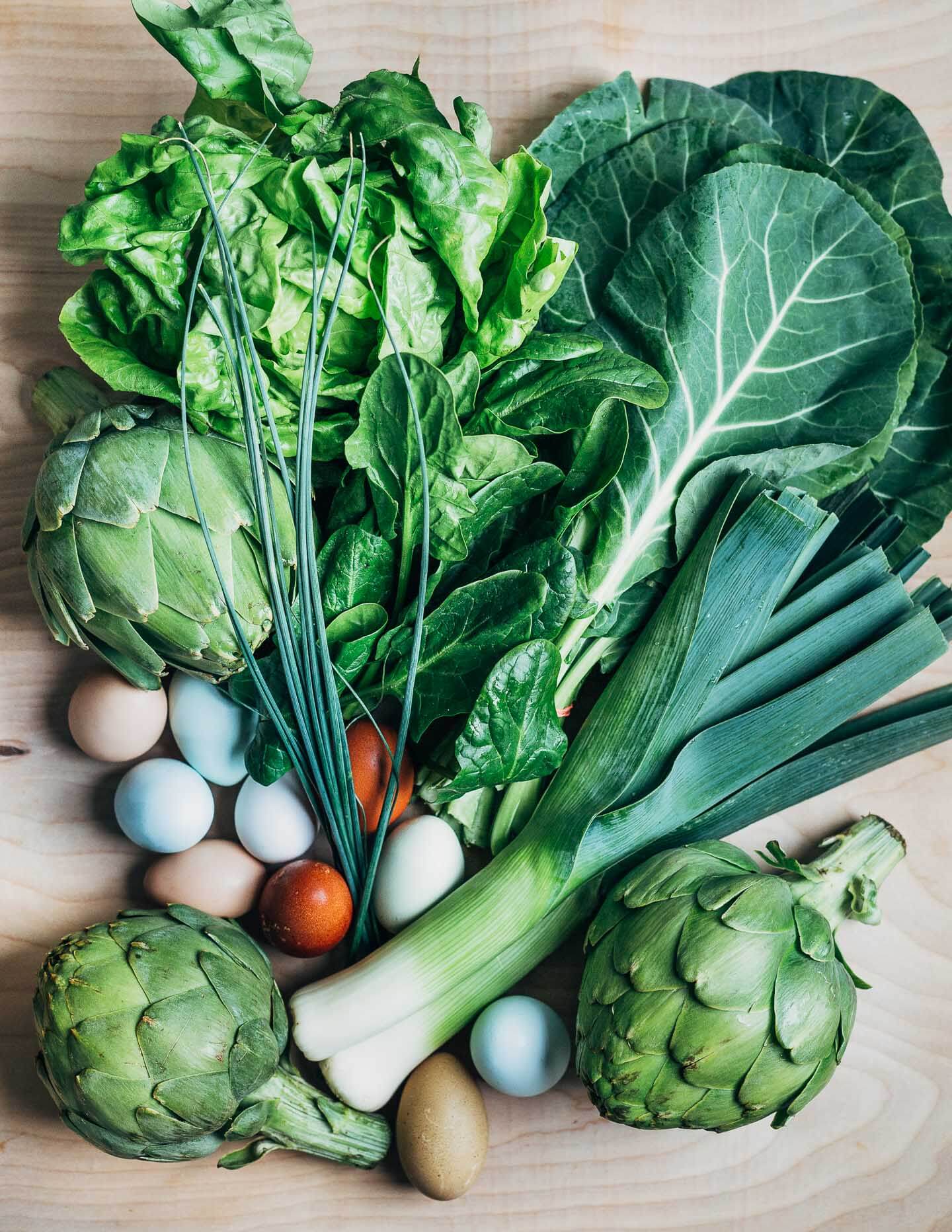 Produce laid out on a table top, with artichokes, leeks, blue and brown eggs, chives, collards, and lettuce. 