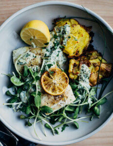 A bowl with lemon wedges, cod, potatoes, micro greens and a creamy sauce.