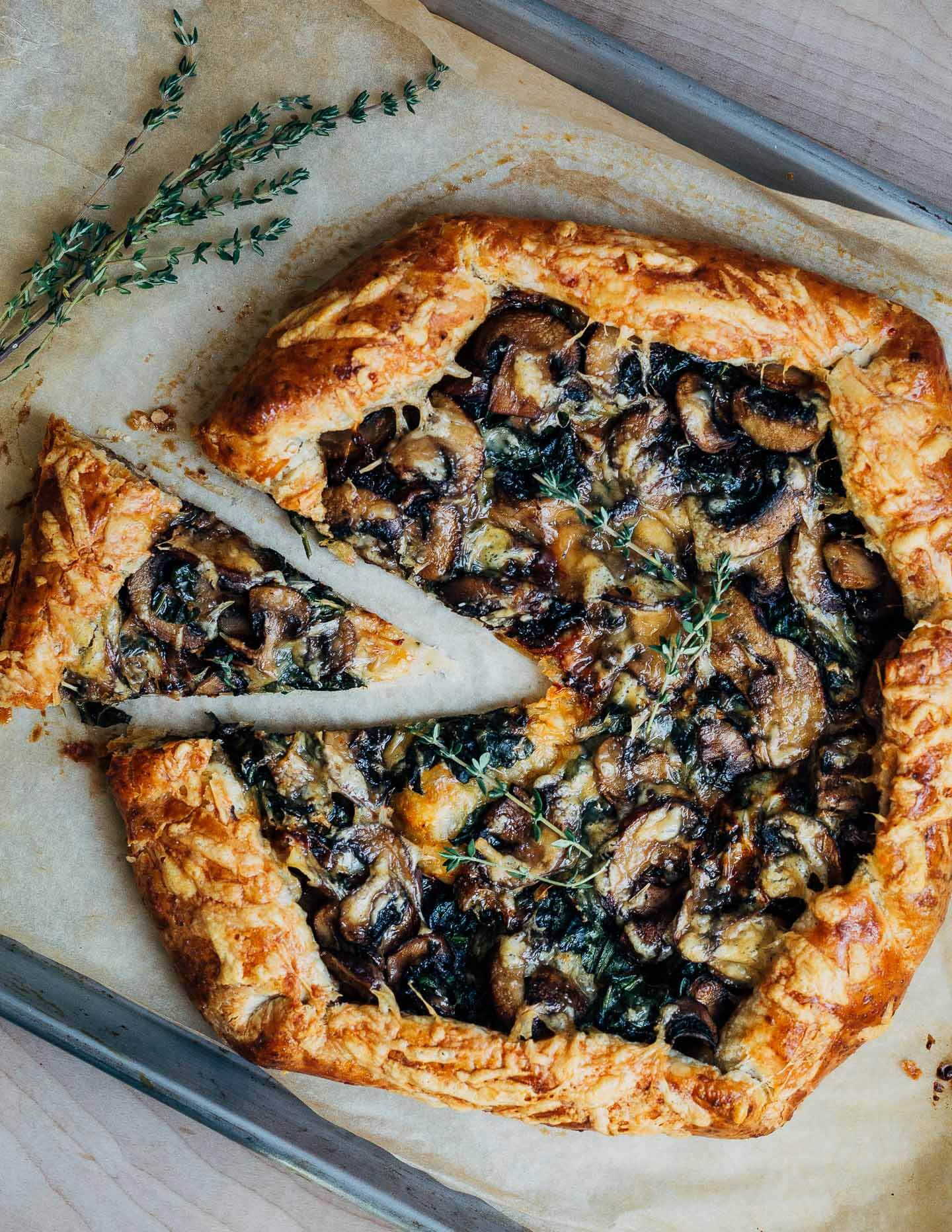 A mushroom galette on a parchment-lined baking sheet with one slice cut and some fresh thyme alongside. 