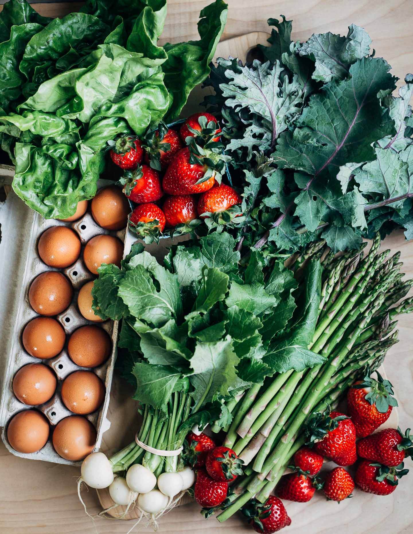 A tabletop with piles of lettuce, kale, asparagus, strawberries, and brown eggs. 