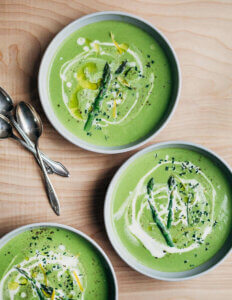 Three bowls of asparagus soup with swirls of cream and olive oil, fresh herbs, and asparagus tips.