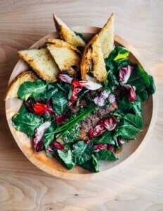 A bowl of kale salad topped with sliced steak, golden croutons, and herbs.