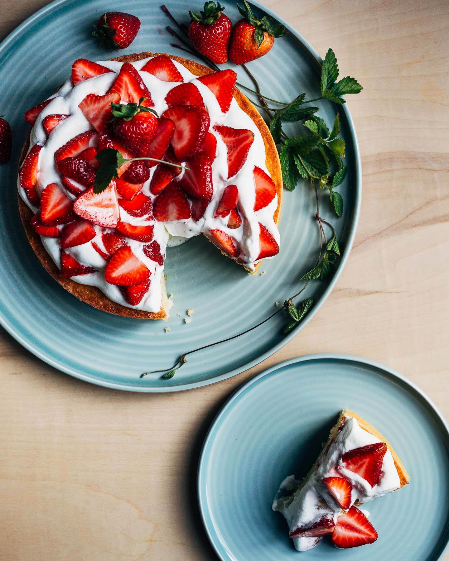 An overhead view of a cake topped with strawberries and cream, with a slice on a plate alongside. 