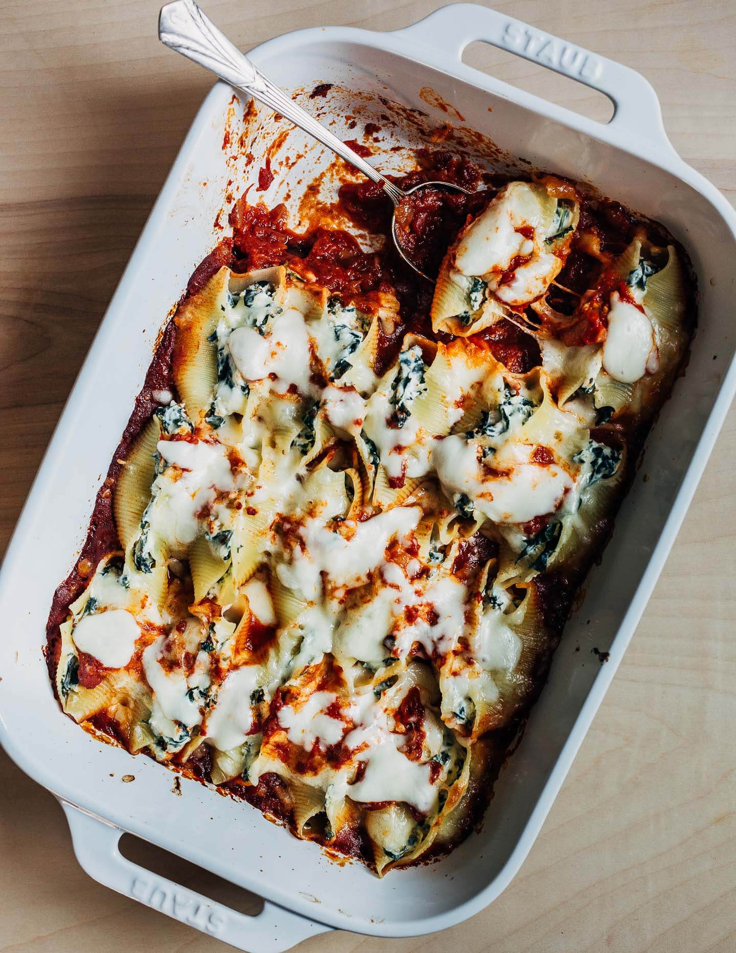 A baking dish with cheese-covered stuffed shells. A spoon is in the dish and a few shells have already been served. 