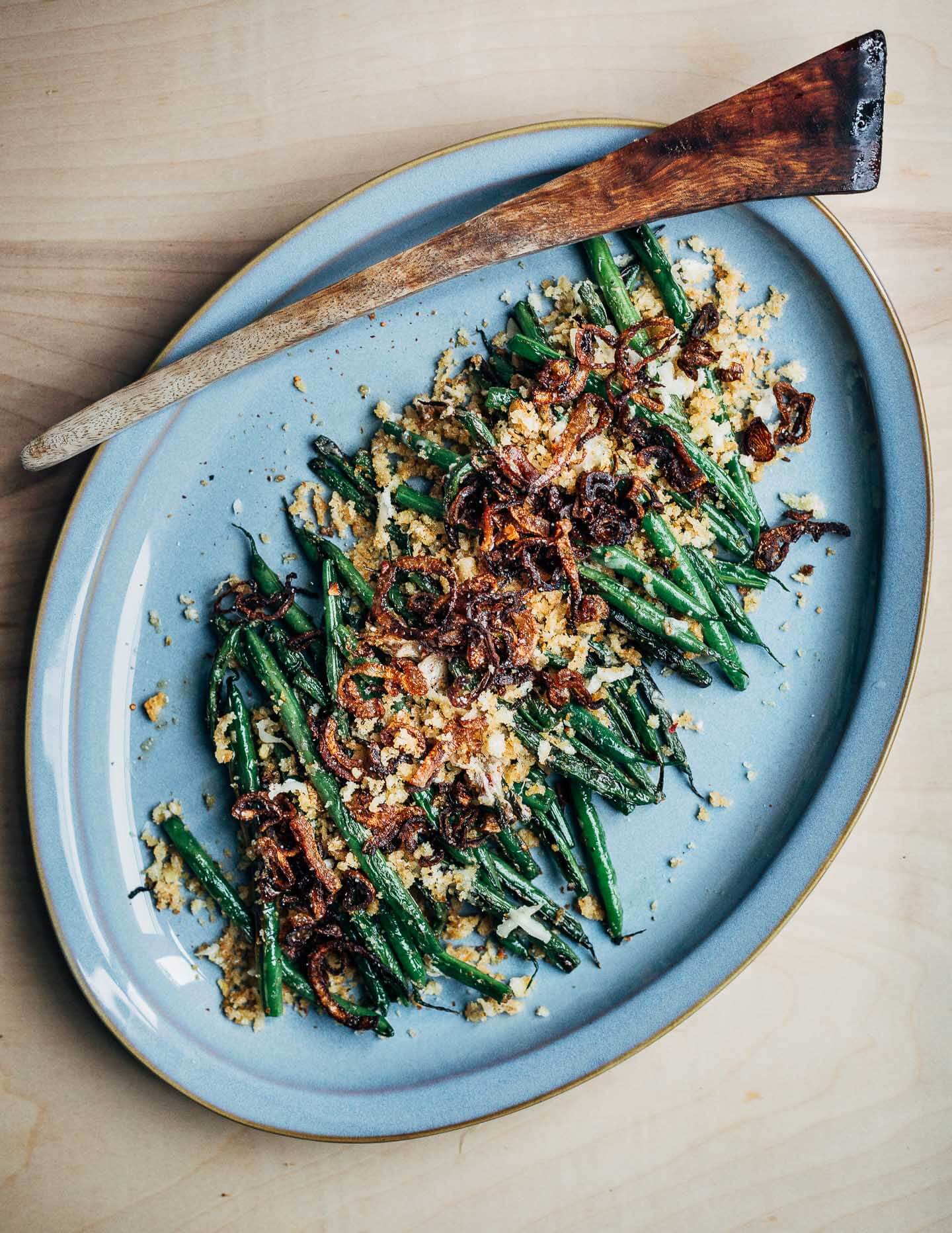 A platter of cooked green beans topped with golden breadcrumbs and fried shallots.