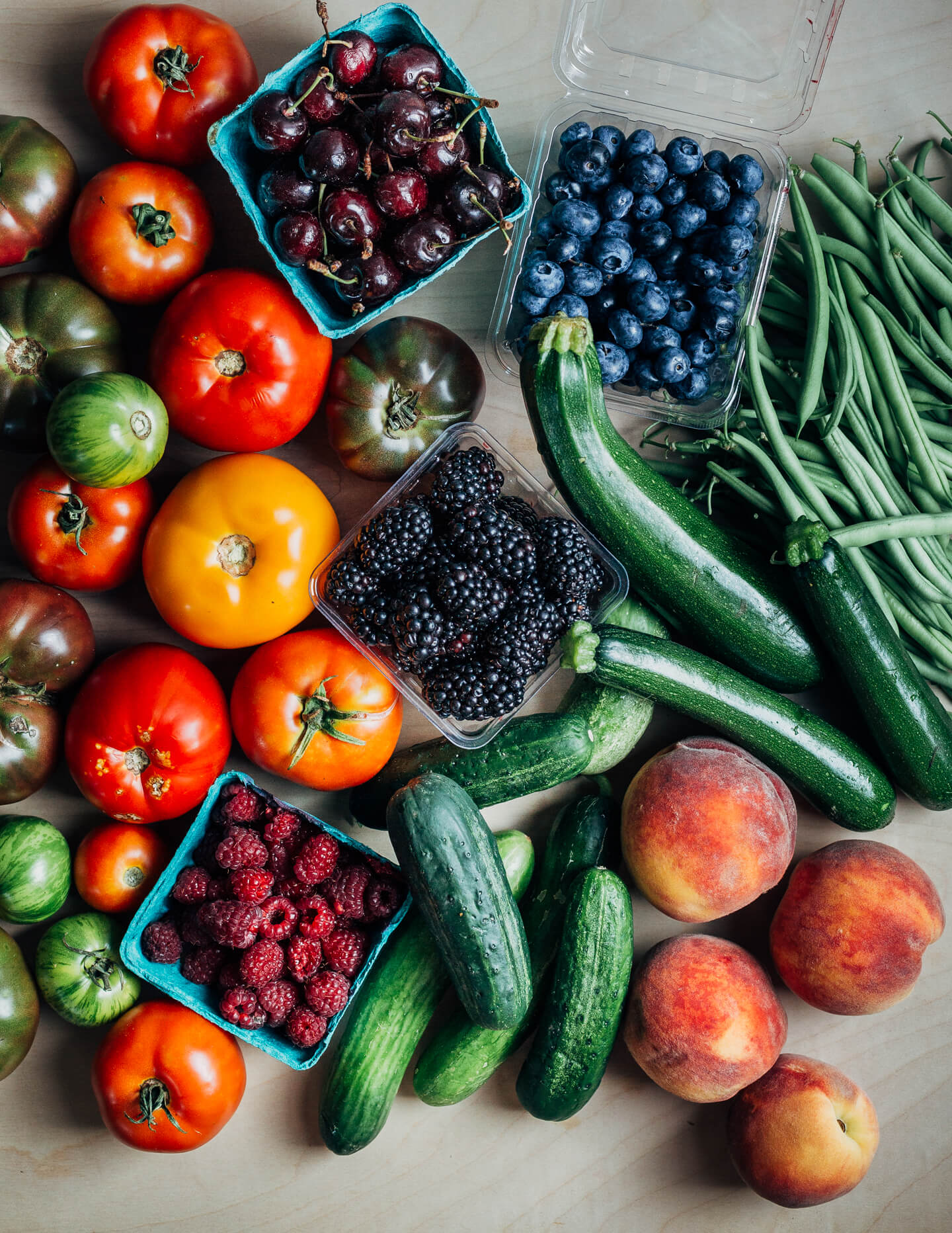 A table covered with fruit and produce, including tomatoes, berries and cherries, cucumber, zucchini, peaches, and green beans. 