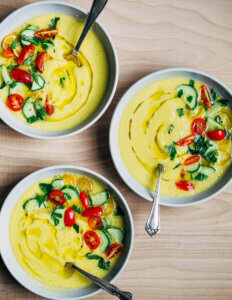 Three bowls of yellow gazpacho garnished with cucumbers and tomatoes.