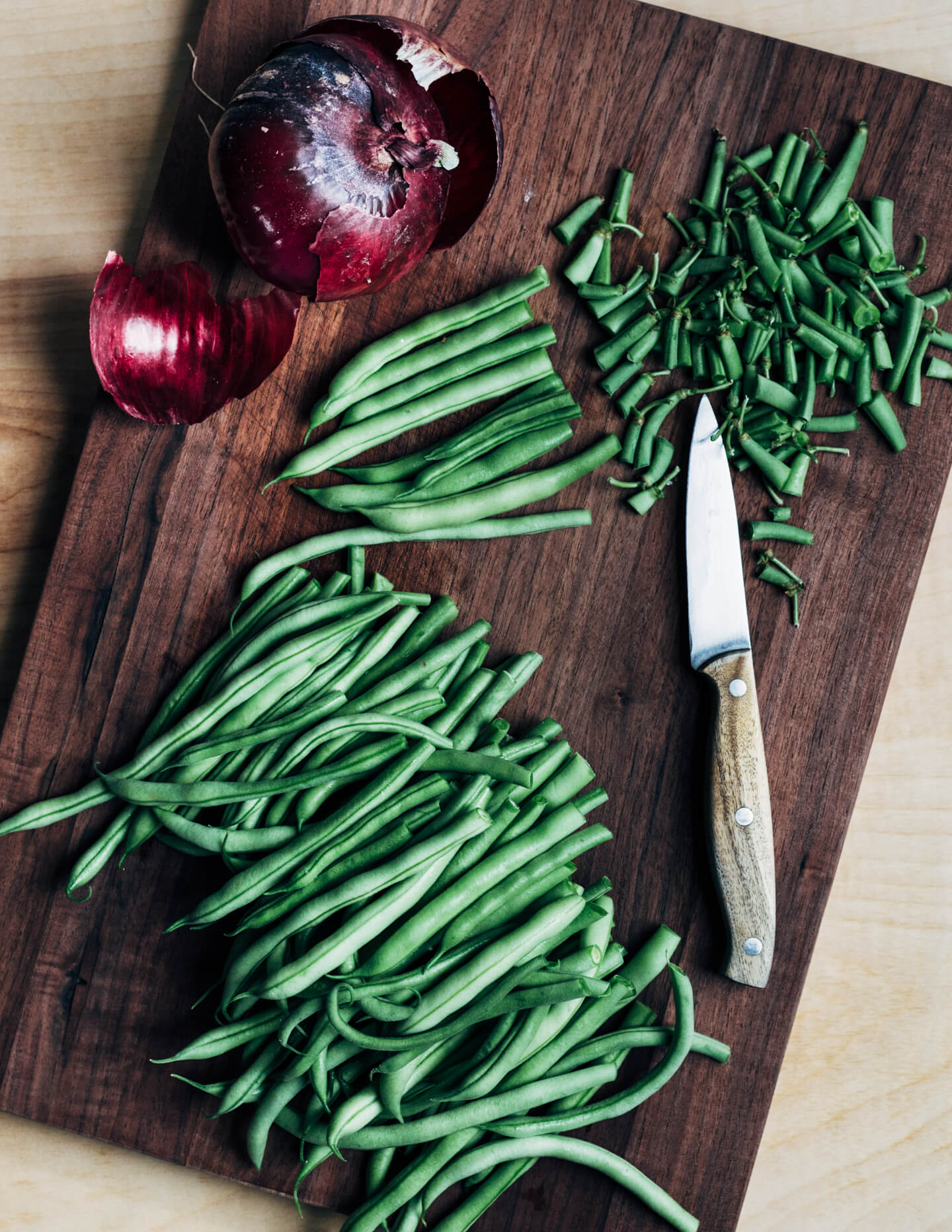 A cutting board with a knife trimming green beans and a red onion. 