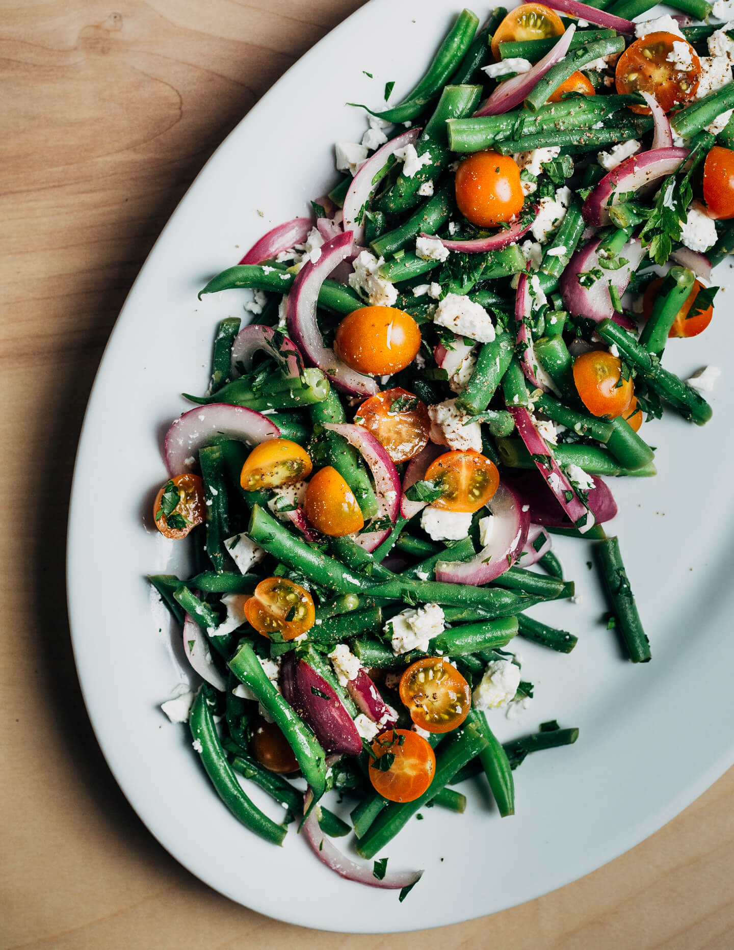 A white platter with green bean salad, with red onions, tomatoes, and feta.