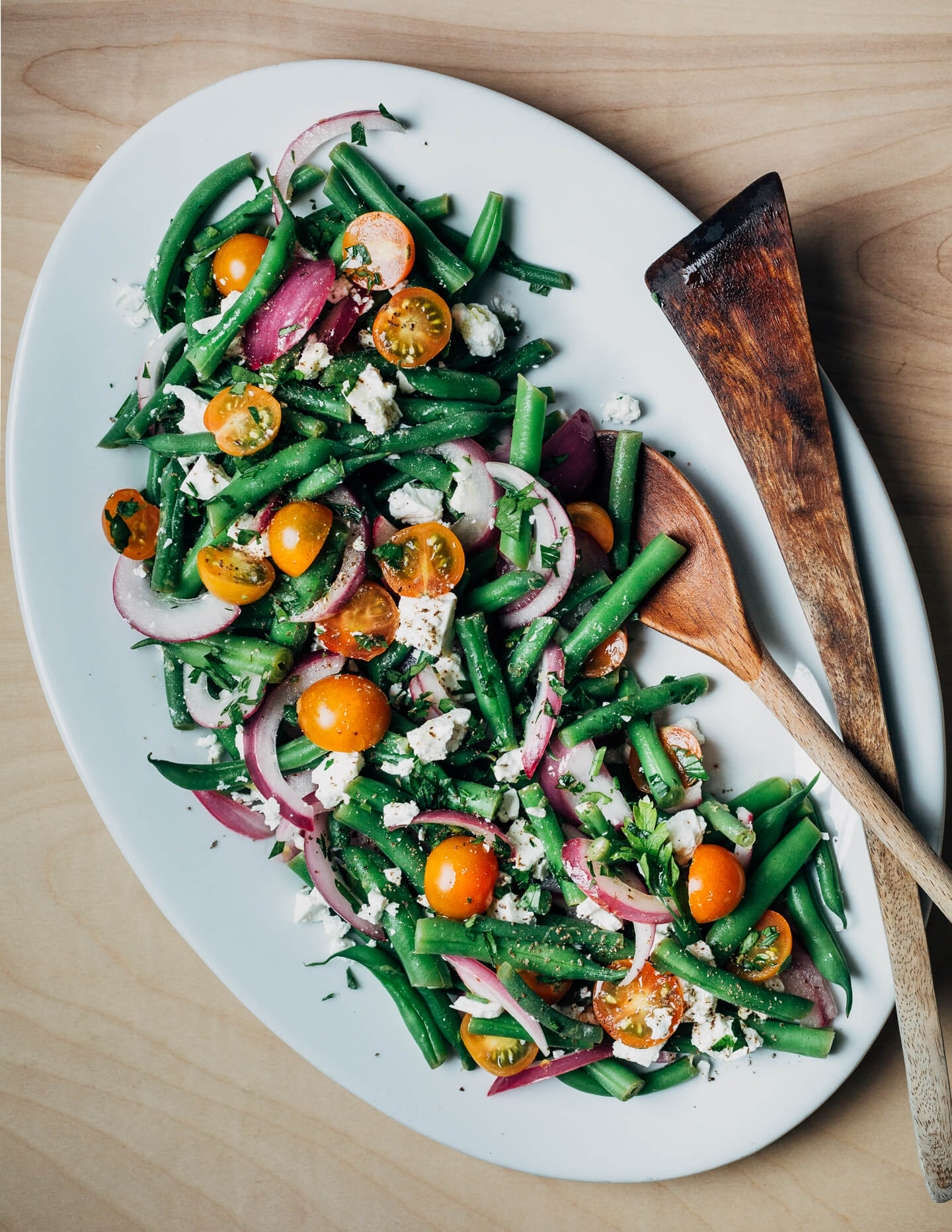 A white platter with green bean salad, with red onions, tomatoes, and feta. There are wooden serving utensils on the side. 