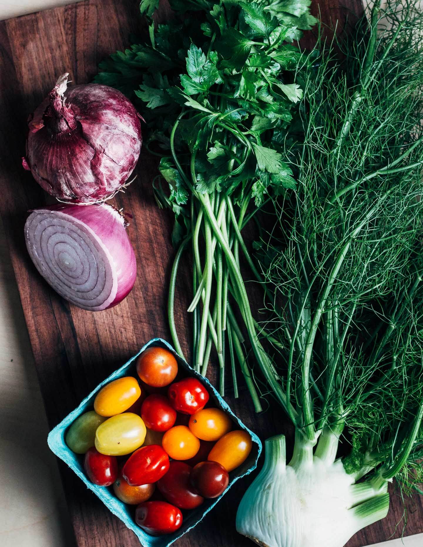 A cutting board with cherry tomatoes, fennel, red onions, and parsley. 