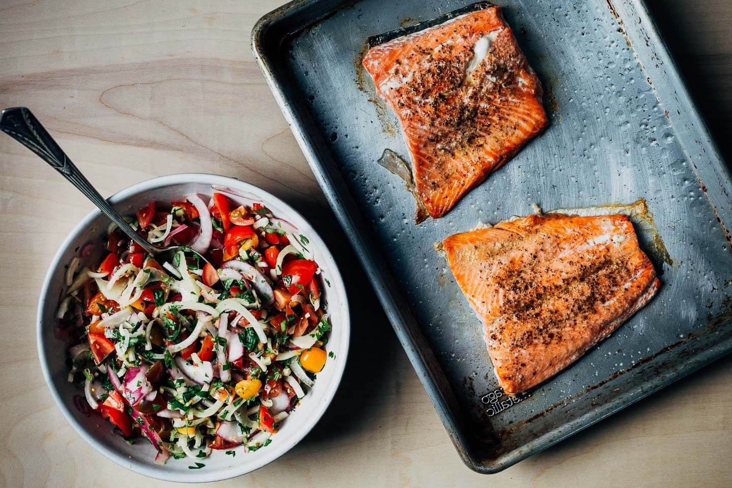 A bowl of tomato-fennel salad alongside a tray of broiled salmon filets. 