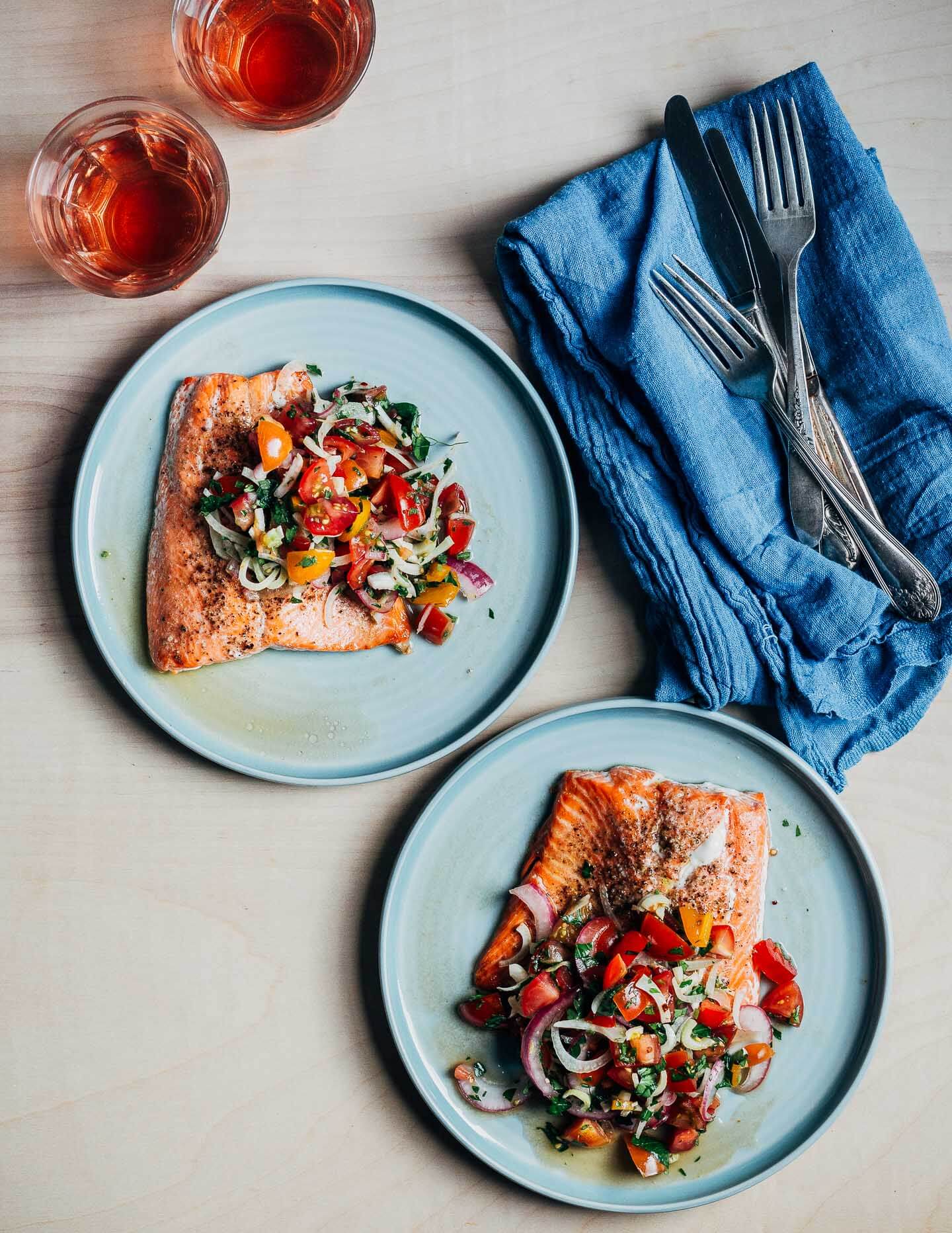 Glasses of rosé wine, two plates with salmon filets and tomato salad, and an indigo napkin alongside. 