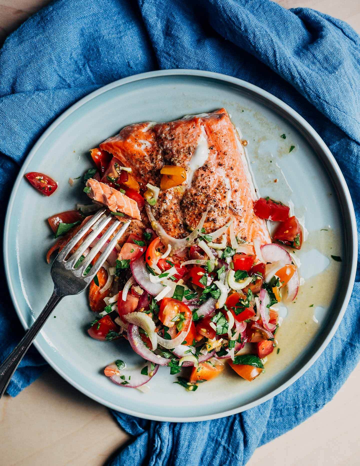 A fork taking a bite out of a salmon filet topped with a tomato salad. 