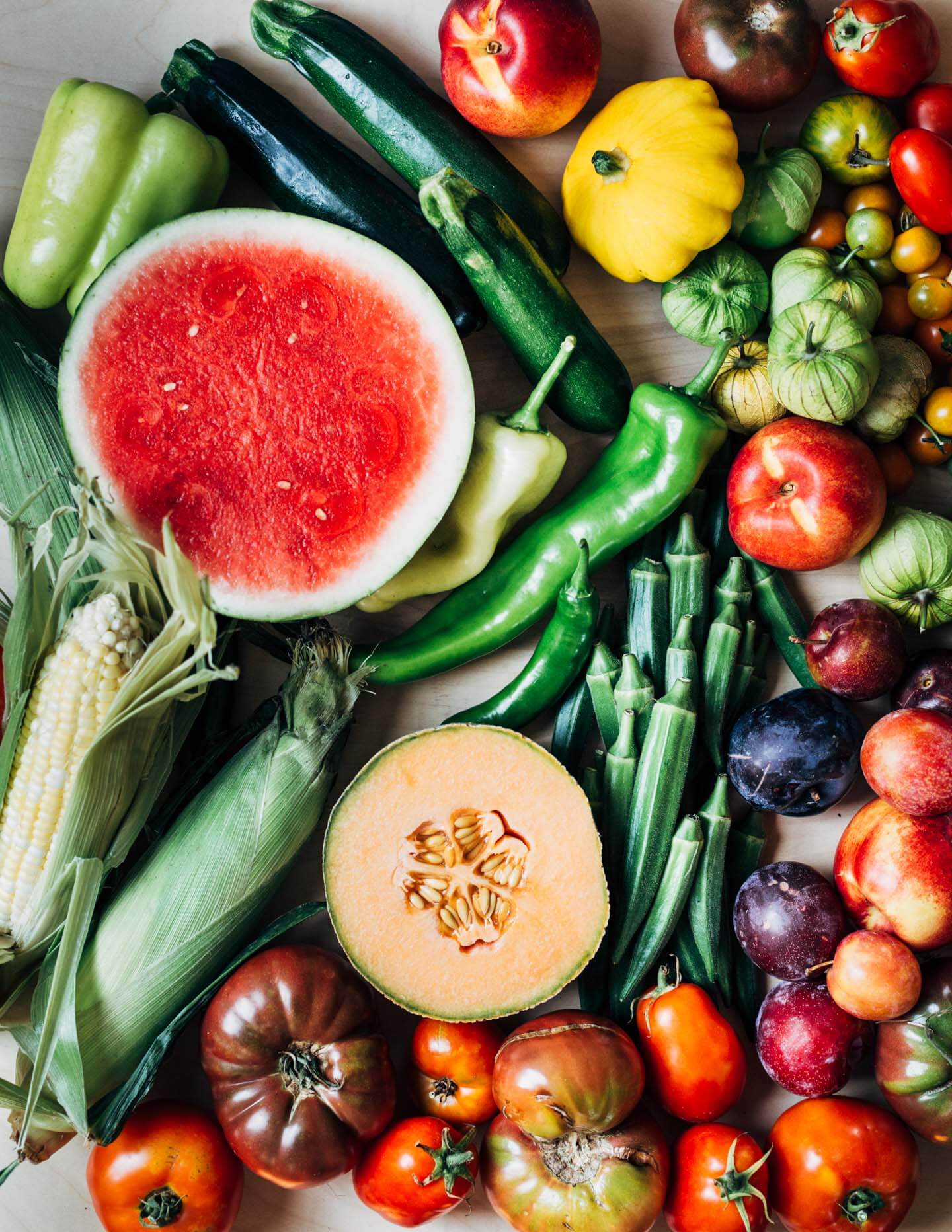 A tabletop covered in colorful fruits and vegetables, including watermelon, cantaloupe, tomatoes, tomatillos, oka, corn, and zucchini. 