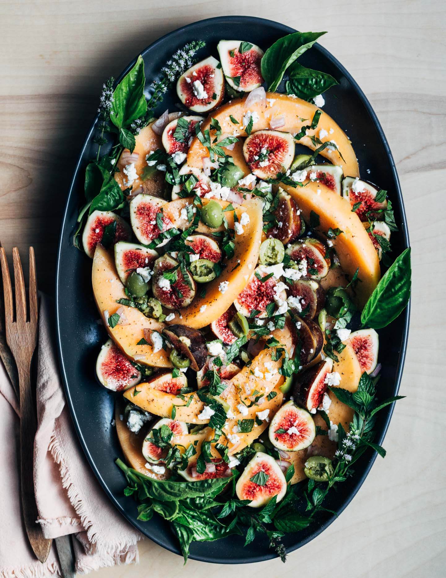 A large platter with cantaloupe, figs, feta, olives, and herbs.