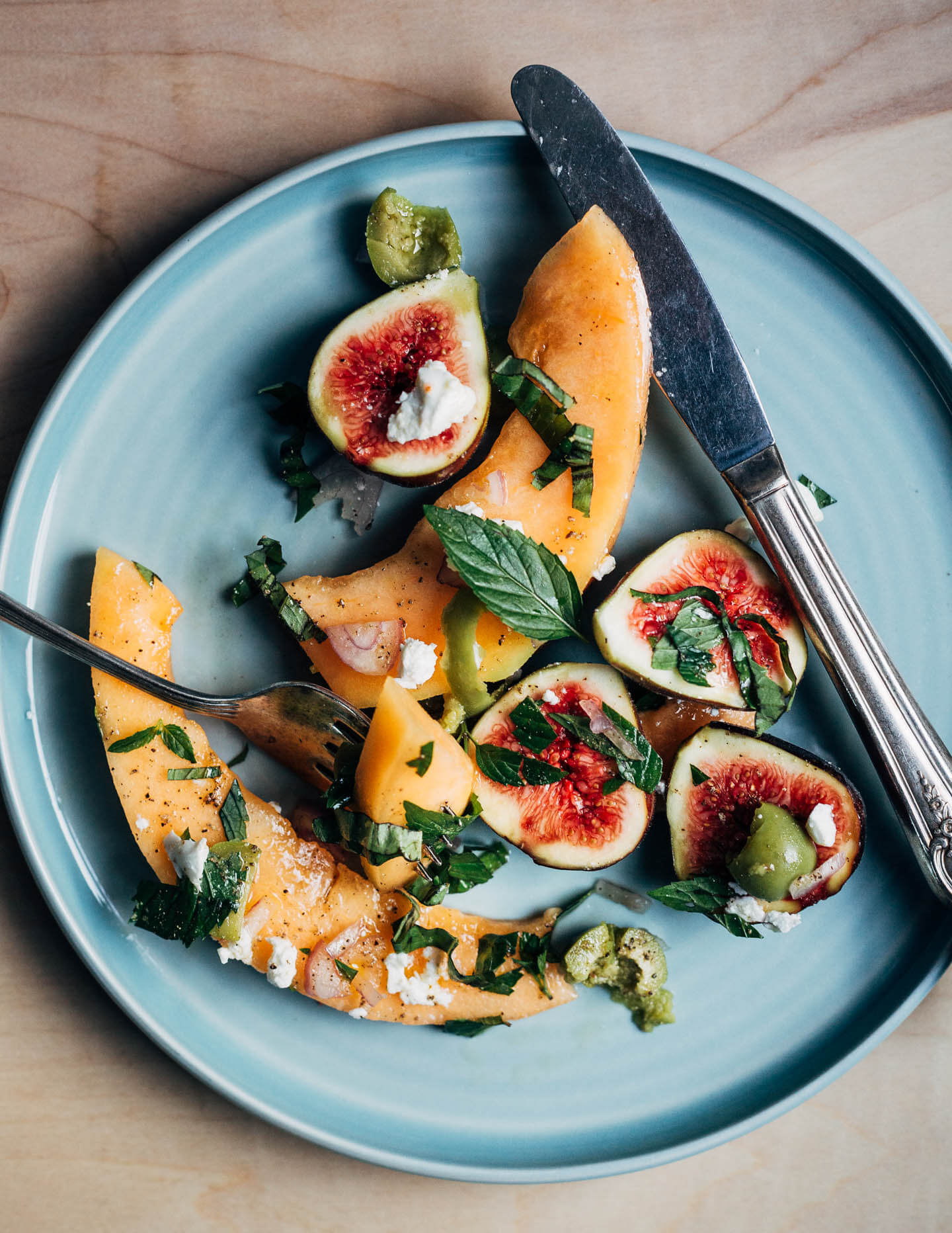 A plate with cantaloupe wedges, figs, feta, and herbs. 