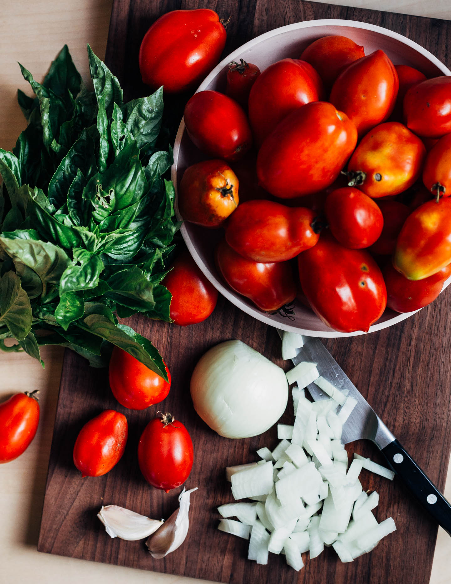 A cutting board with chopped onions, garlic cloves, basil, and a bowl of Roma tomatoes. 