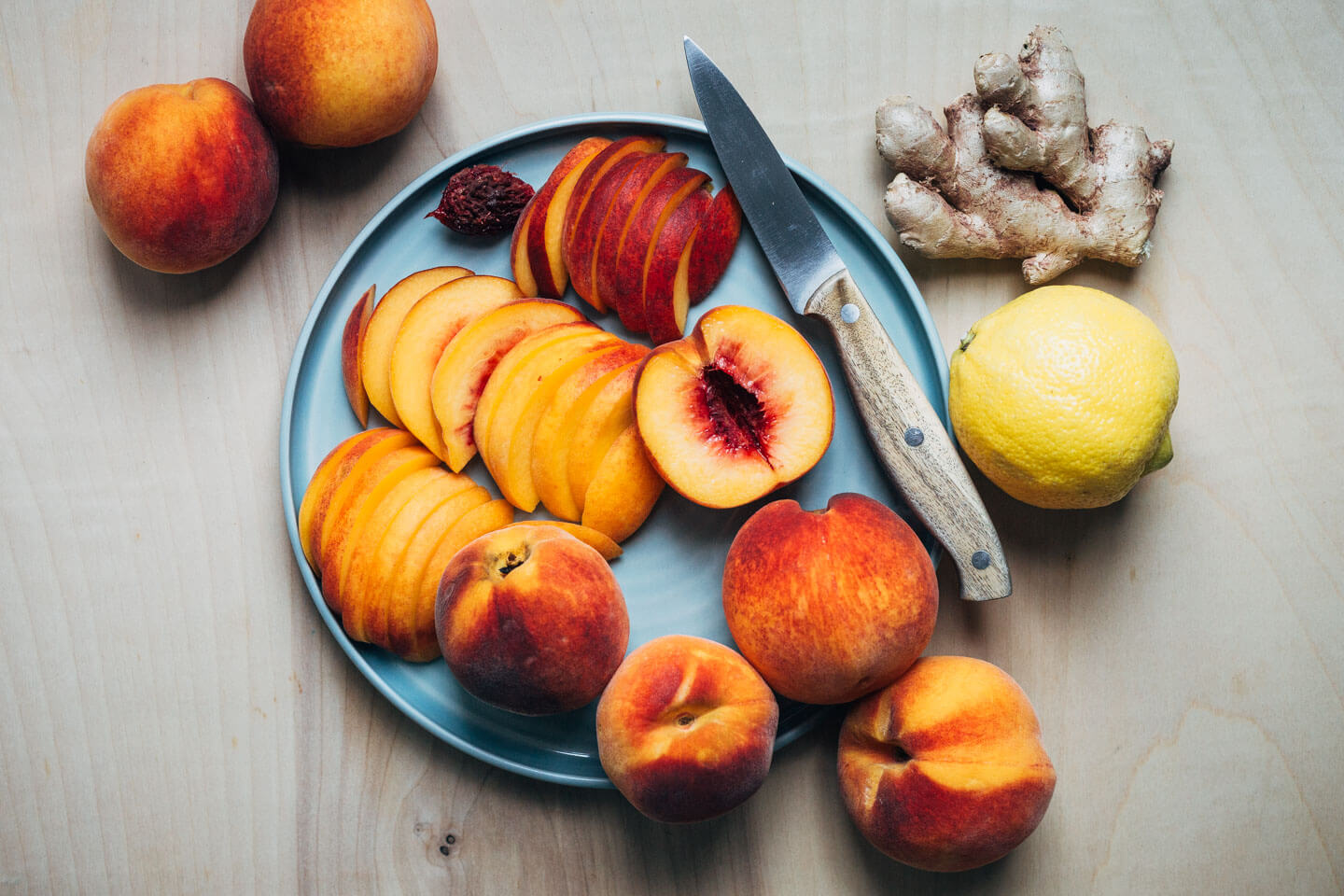 A plate with sliced peaches and a paring knife, plus ginger, lemon, and whole peaches alongside.