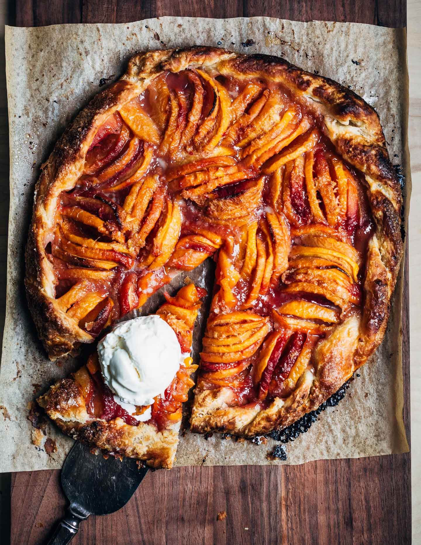 A peach galette on a wooden board with a slice cut out. The slice has a scoop of vanilla ice cream on top. 