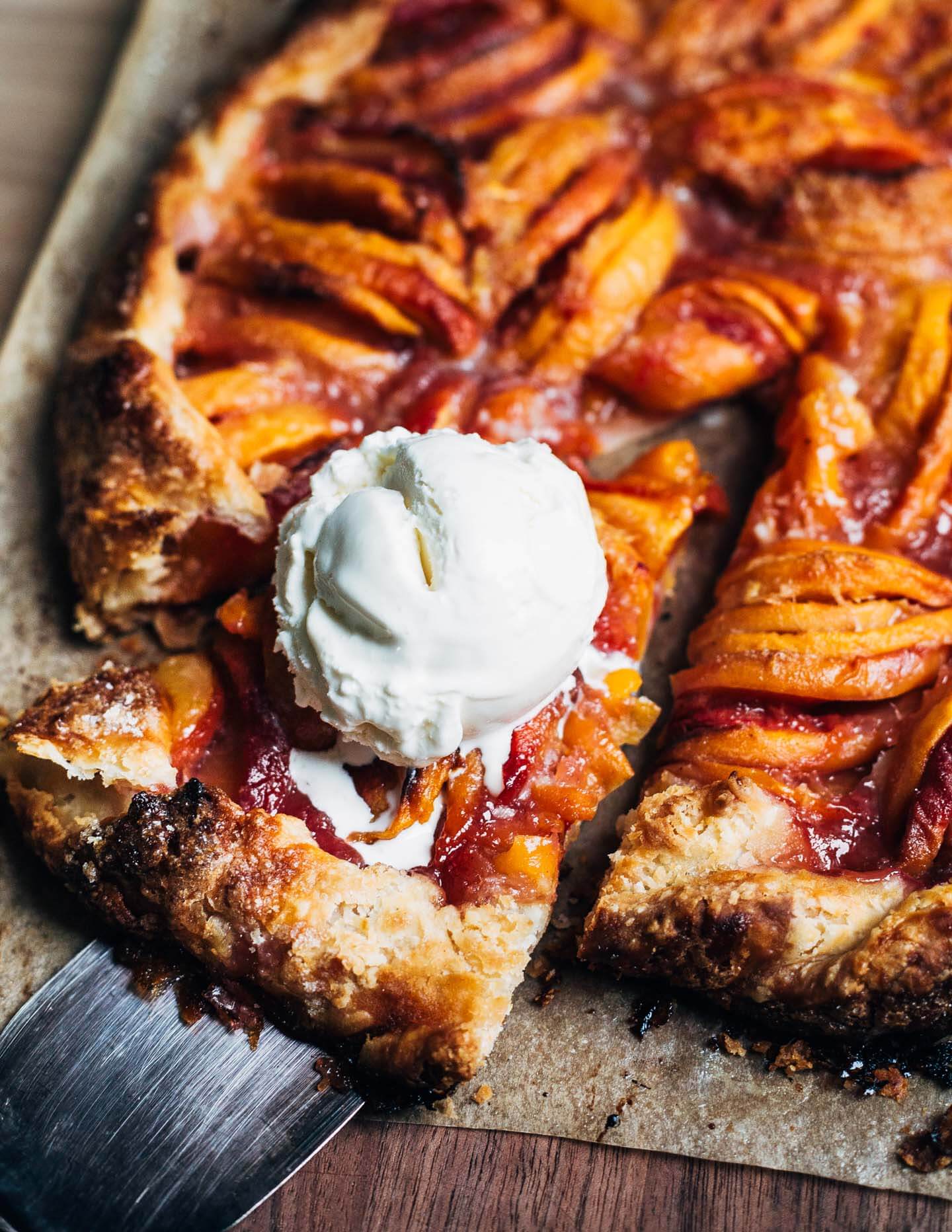 A peach galette on a wooden board with a slice cut out. The slice has a scoop of vanilla ice cream on top.