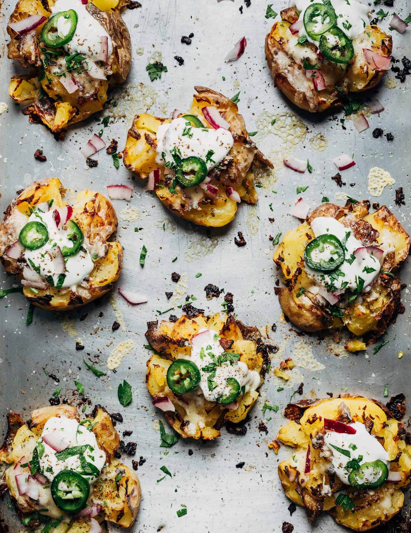 A sheet pan with smashed potatoes topped with sour cream, jalapeños, red onions, and herbs. 