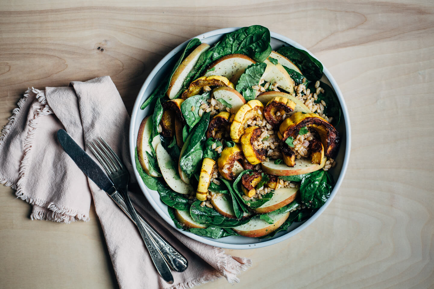 A wide bowl with greens, roasted squash, apples, and farro. A napkin with cutlery is alongside.