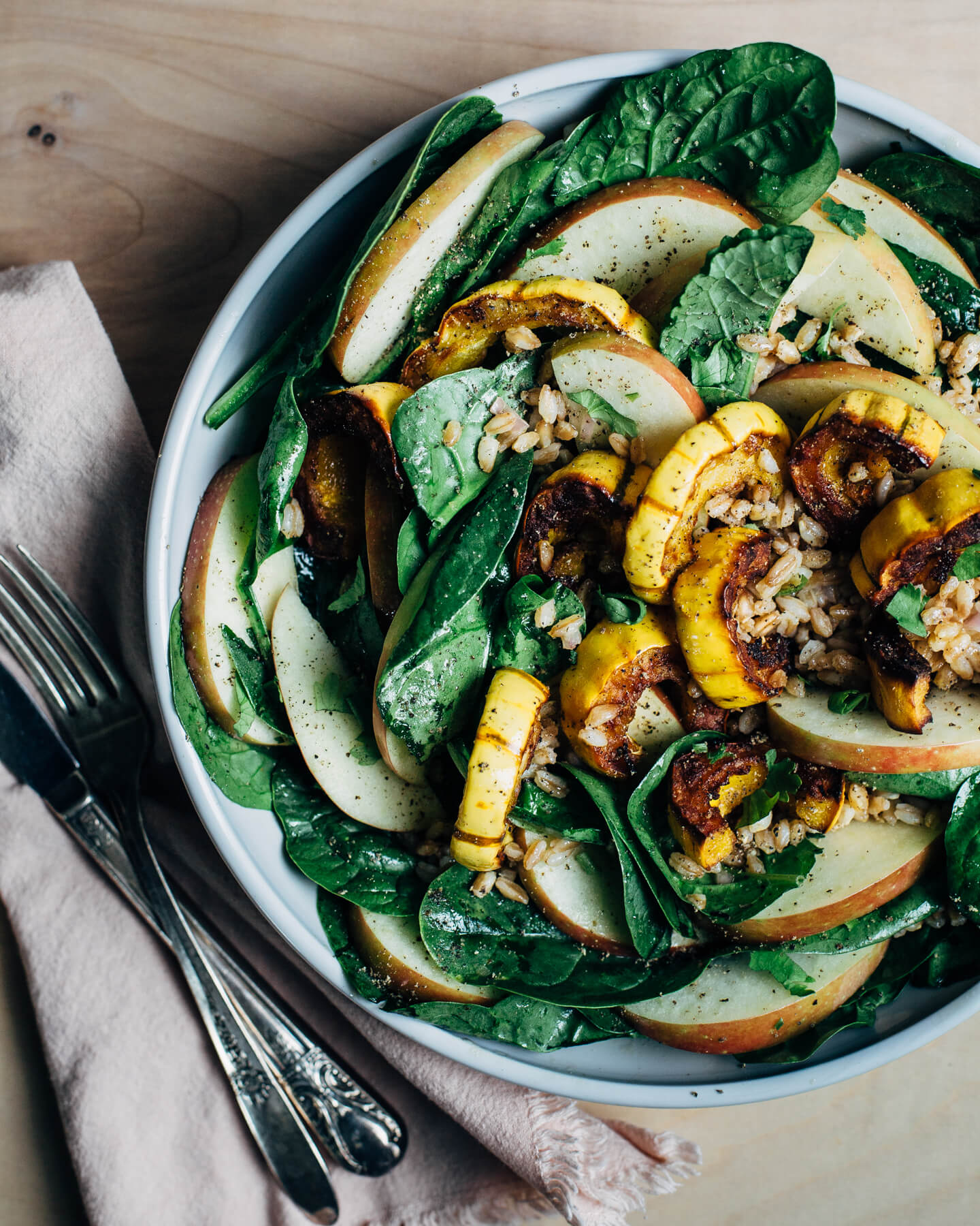 A wide bowl with greens, roasted squash, apples, and farro. A napkin with cutlery is alongside. 