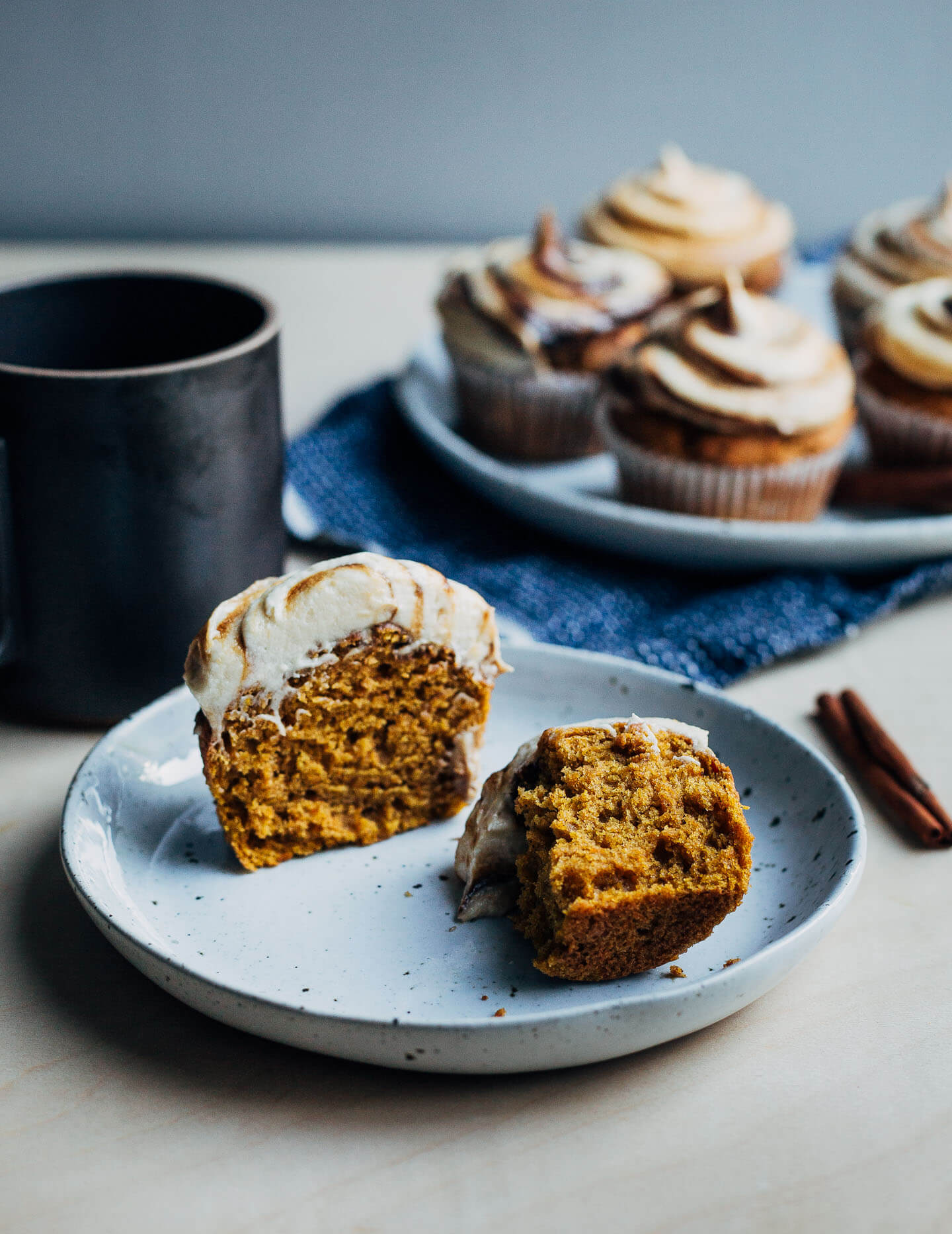 A pumpkin cupcake on a plate, with a mug of tea and a plate of cupcakes in the background. 