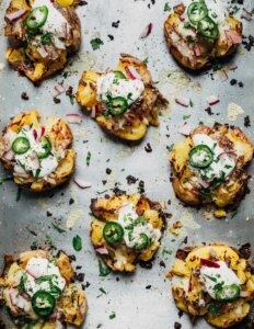 Smashed potatoes on a baking sheet. They are topped with jalapenos, herbs, and sour cream.