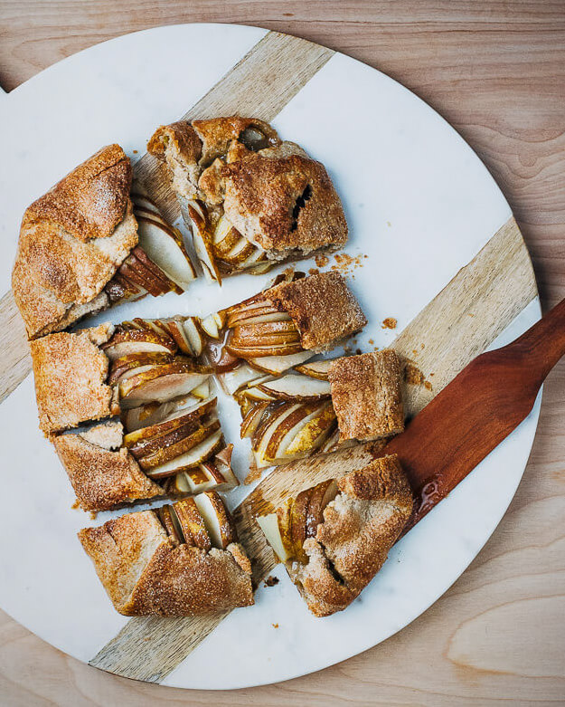 A cut up pear galette on a marble slab with a wooden server alongside. 