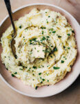 A pink serving bowl with mashed potatoes. The potatoes are dotted with herbs and a pat of butter.