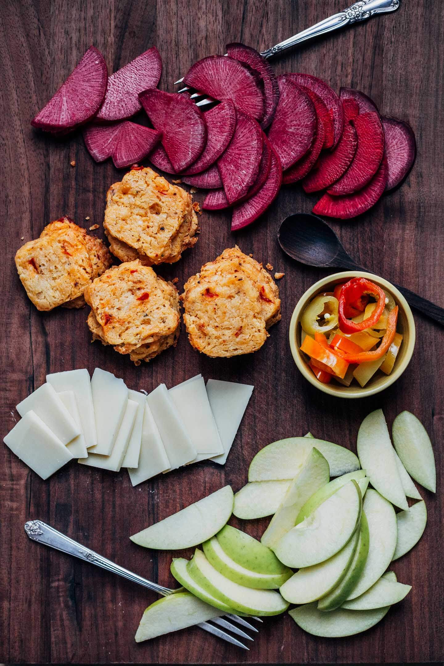 A snack board with radishes, cheese crackers, cheese slices, apples, and peppers. 
