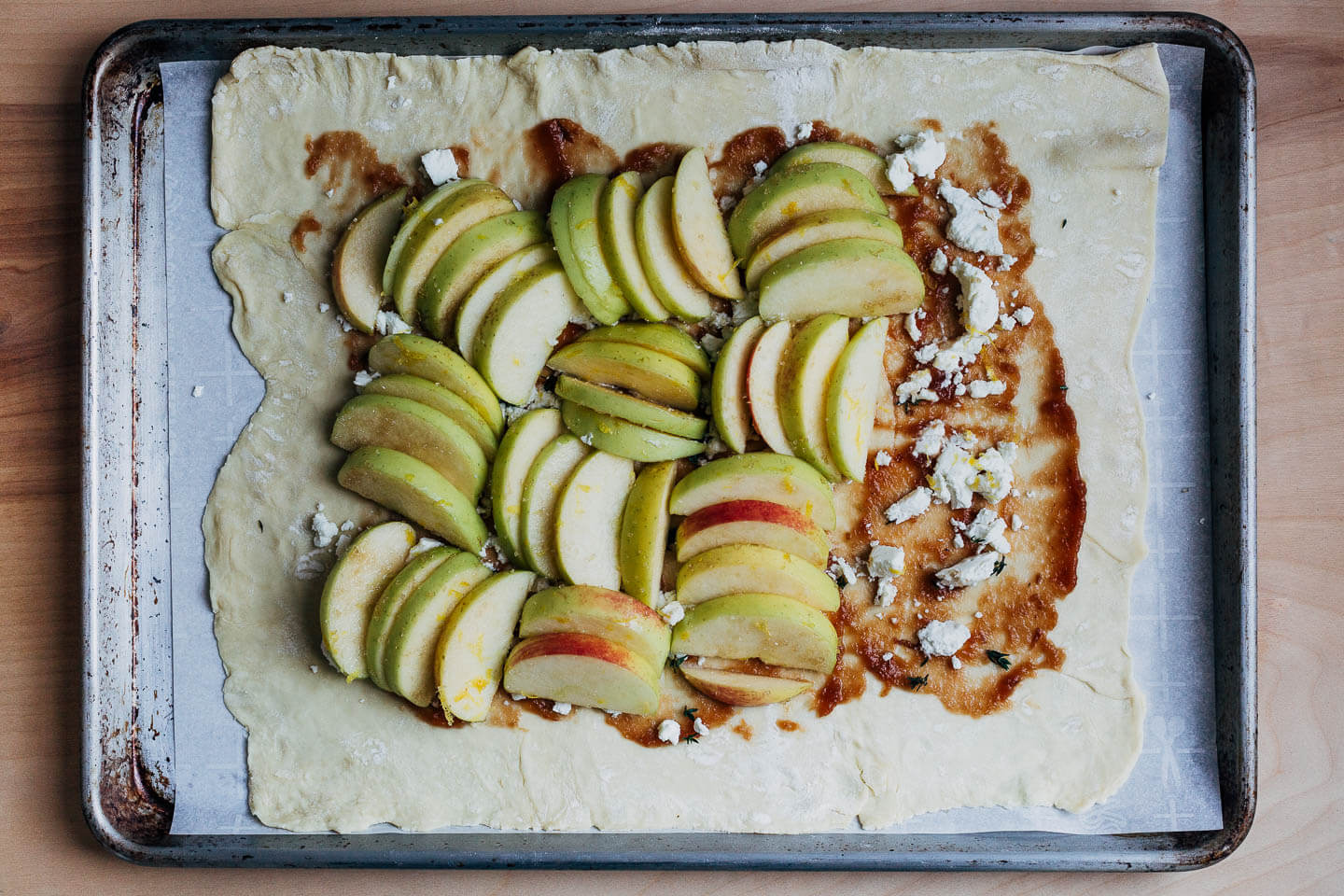Pastry dough on a baking sheet with apple butter, goat cheese, and clusters of apples. 