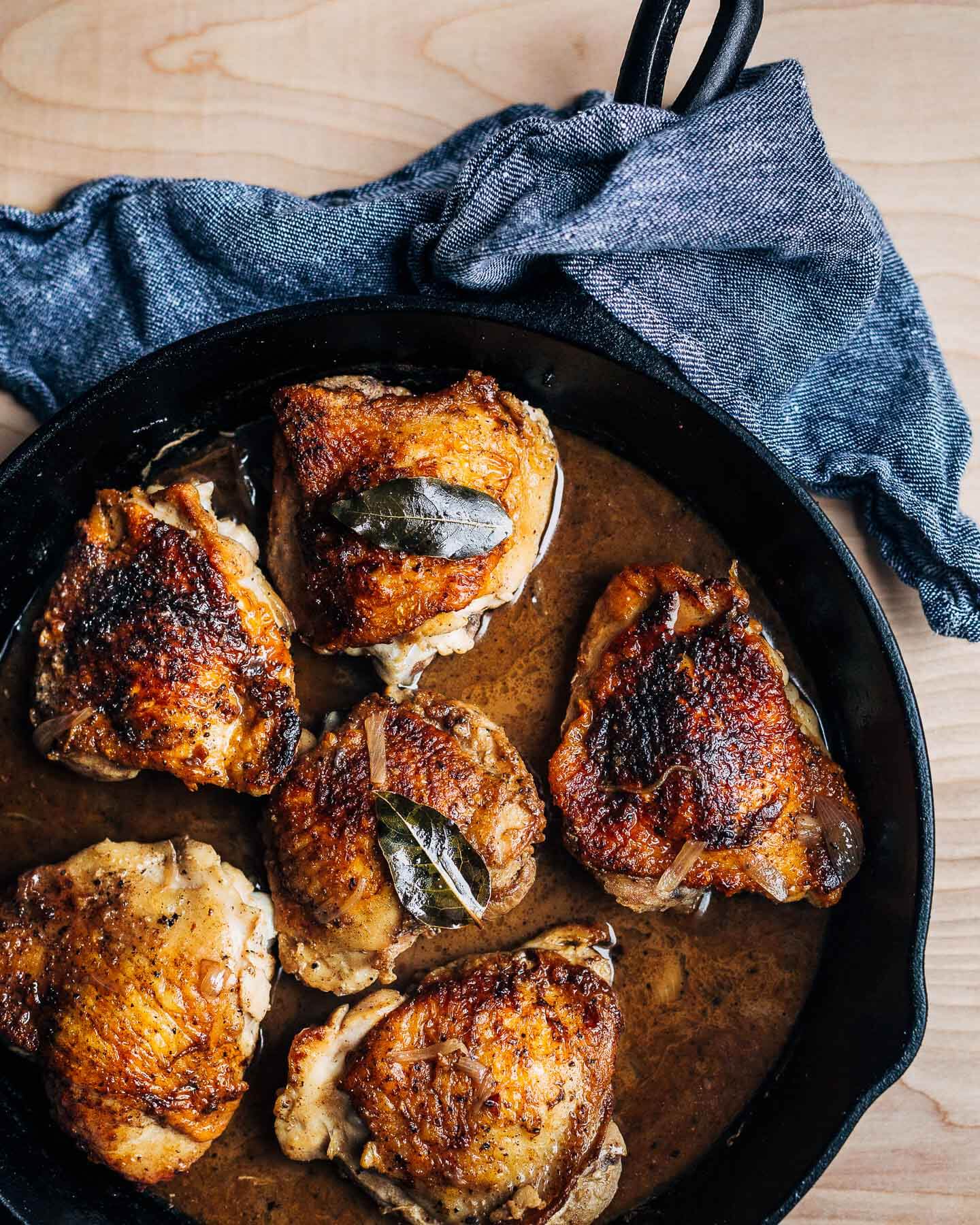 A cast iron skillet of deep golden brown chicken thighs with a bay leaf on top. 