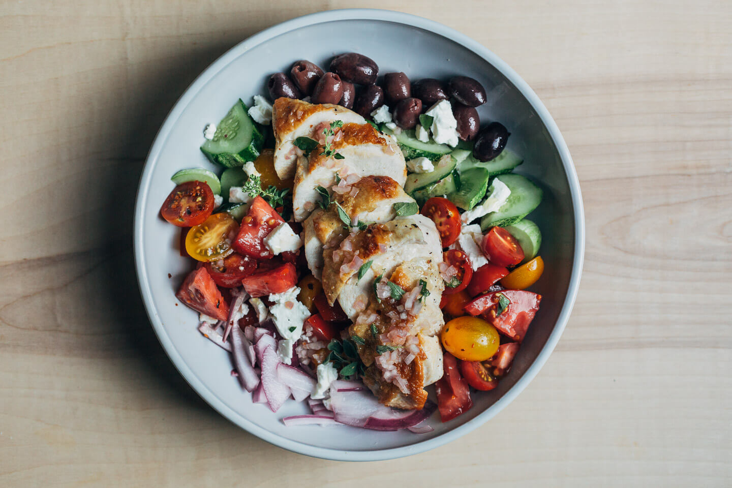 A wide bowl with a Greek salad topped with sliced chicken breasts. 