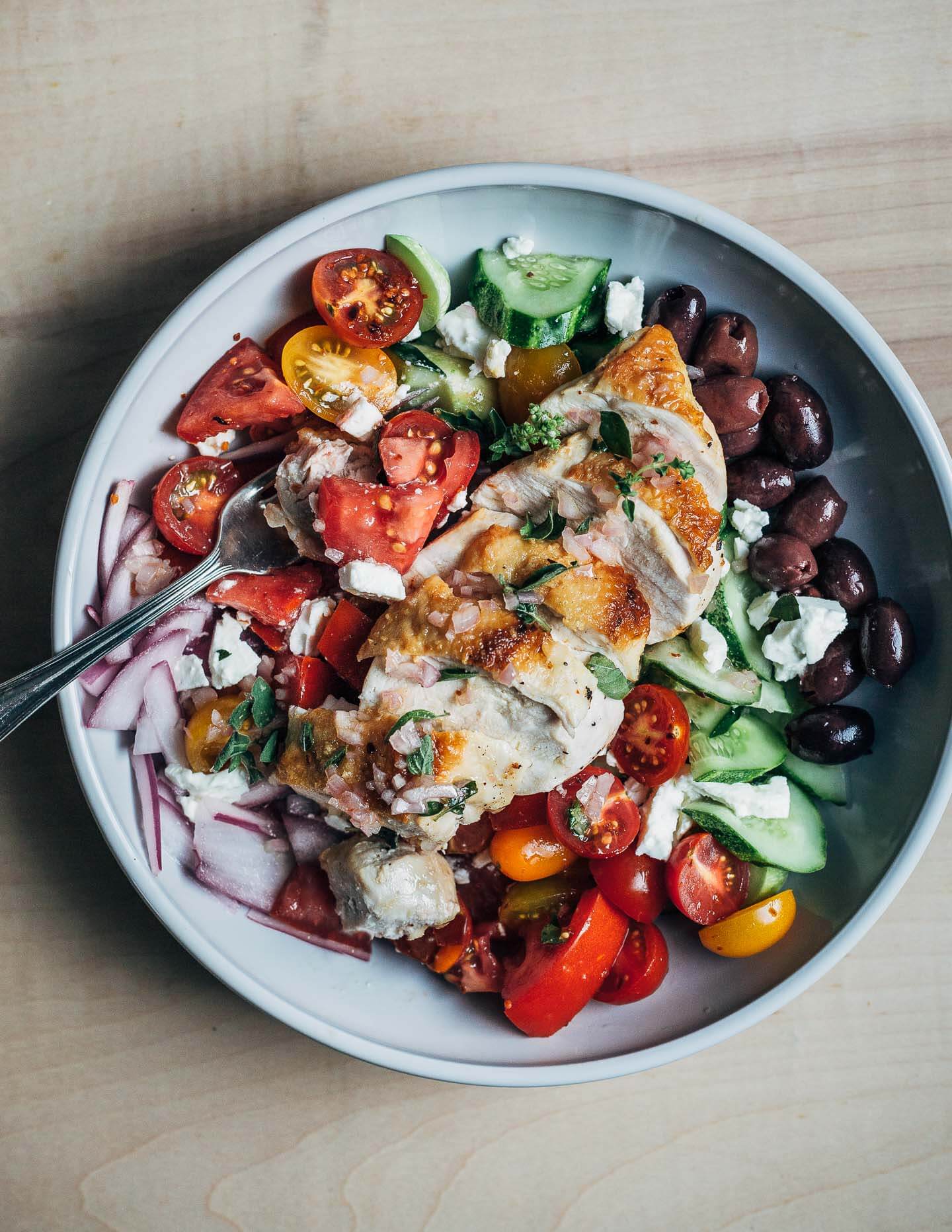A wide bowl with a Greek salad topped with sliced chicken breasts. 